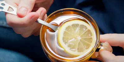 Nahaufnahme von Händen, die ein Glas mit heißem Zitronentee mit Honig und einen Löffel halten.