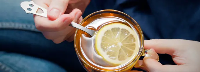 Nahaufnahme von Händen, die ein Glas mit heißem Zitronentee mit Honig und einen Löffel halten.