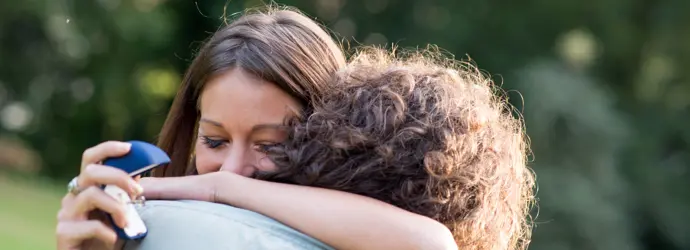 Una ragazza castana abbraccia un ragazzo con i capelli castani ricci e tiene in mano un cofanetto per anello blu.