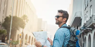 Ein Mann, der einen blau-grauen Rucksack und eine dunkle Sonnenbrille trägt, hält an einer Straßenecke einen Stadtplan.