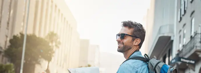 Ein Mann, der einen blau-grauen Rucksack und eine dunkle Sonnenbrille trägt, hält an einer Straßenecke einen Stadtplan.