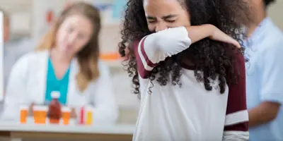 Una ragazza con i capelli ricci castani starnutisce nel gomito. Sullo sfondo due persone sfocate.