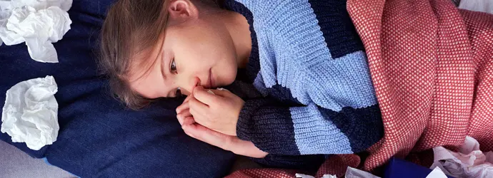 Ein junges Mädchen liegt mit einer Decke zugedeckt auf einem Sofa und ist umgeben von benutzten Taschentüchern.