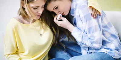 Una ragazza castana con camicia a quadri azzurra si pulisce il naso con un fazzoletto mentre la sua amica la abbraccia.