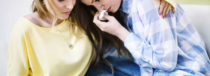 Una ragazza castana con camicia a quadri azzurra si pulisce il naso con un fazzoletto mentre la sua amica la abbraccia.