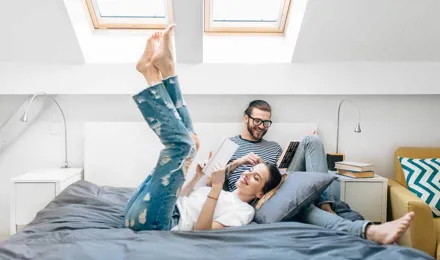 Ein junges Paar sitzt lesend auf einem Bett in einem Zimmer mit zwei Deckenleuchten