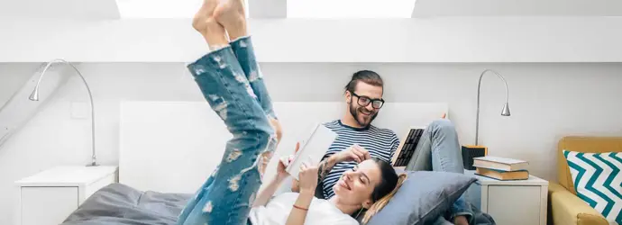 Ein junges Paar sitzt lesend auf einem Bett in einem Zimmer mit zwei Deckenleuchten