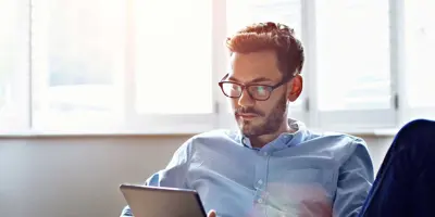 Ein Mann mit Brille sitzt auf einem Sofa und blickt auf ein digitales Tablet