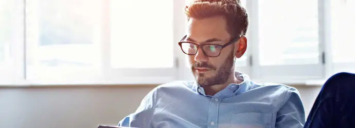 Ein Mann mit Brille sitzt auf einem Sofa und blickt auf ein digitales Tablet