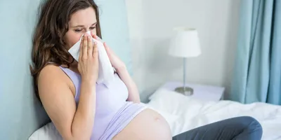 Een zwangere vrouw zit op een bed en snuit haar neus in een zakdoekje