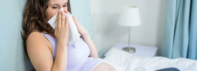 Een zwangere vrouw zit op een bed en snuit haar neus in een zakdoekje