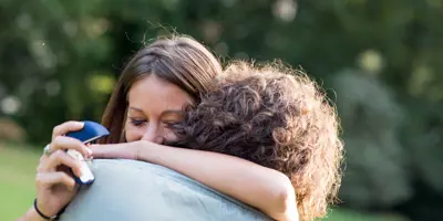 2 personen omarmen elkaar in een tuin terwijl een van hen het doosje van een trouwring vasthoudt