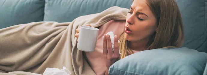 Eine Frau liegt hustend auf dem Sofa und hält eine Teetasse und Taschentücher in den Händen