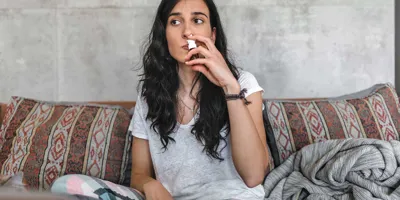 Eine junge Frau mit Birkenpollenallergie sitzt auf einem Sofa und benutzt ein Nasenspray