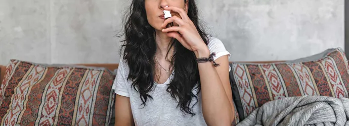 Eine junge Frau mit Birkenpollenallergie sitzt auf einem Sofa und benutzt ein Nasenspray