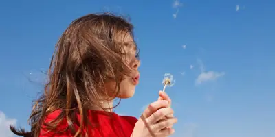 Een jong kind zou huismiddeltjes voor hooikoorts nodig kunnen hebben terwijl ze een paardenbloem blaast met een blauwe lucht op de achtergrond