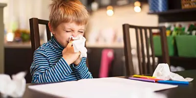 Een kleine jongen snuit zijn neus terwijl hij aan tafel zit met gebruikte tissues om hem heen