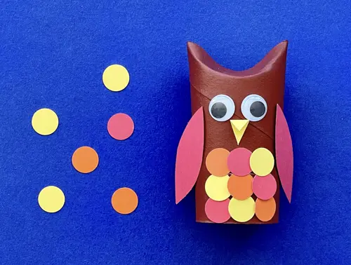 Rotolo di carta a forma di gufo marrone con occhi, becco, ali e cerchi di carta colorati come penne sul petto.