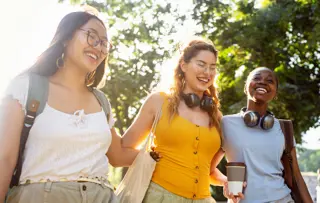 Tre amiche passeggiano all'aperto abbracciate e sorridenti, godendosi il sole.