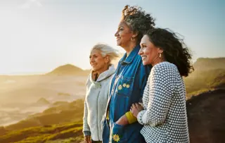 Tre donne in piedi insieme su una collina al tramonto, sorridono e si godono il panorama.
