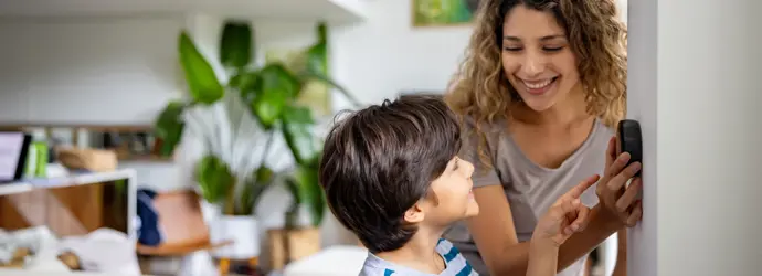 Una donna dai capelli ricci mostra a un ragazzino sorridente un termostato intelligente.