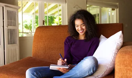 Una ragazza mora e riccia con maglione viola siede su un divano tenendo la testa bassa e sorride.