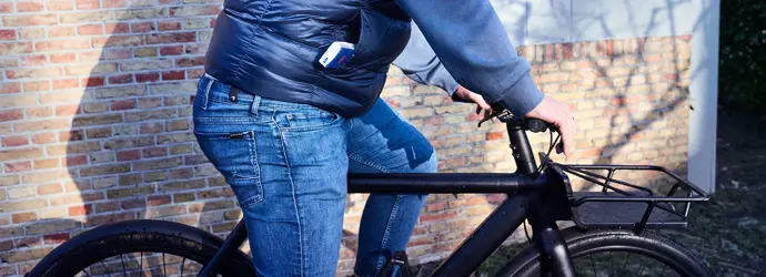 Ein junger Mann in Jeans, Hemd und Weste fährt mit einem schwarzen Fahrrad an einer Steinmauer vorbei.