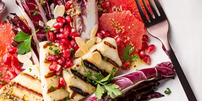 Grillkäse, Radicchio, Granatapfelkerne, Mandelplättchen und Petersilie liegen angerichtet auf einem weißen Teller neben einer Gabel.