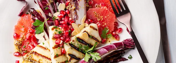 Grillkäse, Radicchio, Granatapfelkerne, Mandelplättchen und Petersilie liegen angerichtet auf einem weißen Teller neben einer Gabel.