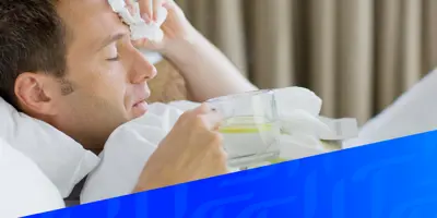 Un uomo è sdraiato a letto e sembra malato, tiene in mano un bicchiere d'acqua e limone e si preme un fazzoletto sulla fronte, con una sovrapposizione geometrica blu in basso nell'immagine.