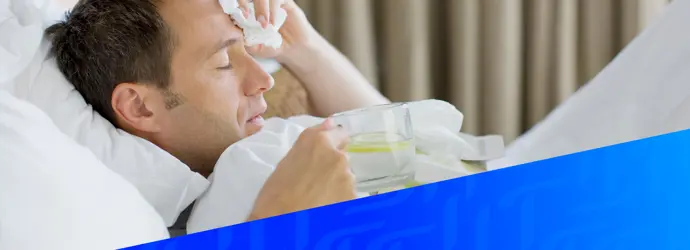 Un uomo è sdraiato a letto e sembra malato, tiene in mano un bicchiere d'acqua e limone e si preme un fazzoletto sulla fronte, con una sovrapposizione geometrica blu in basso nell'immagine.