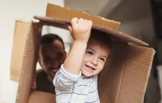 Un bambino sorridente che gioca all'interno di una scatola di cartone mentre il genitore osserva sullo sfondo.