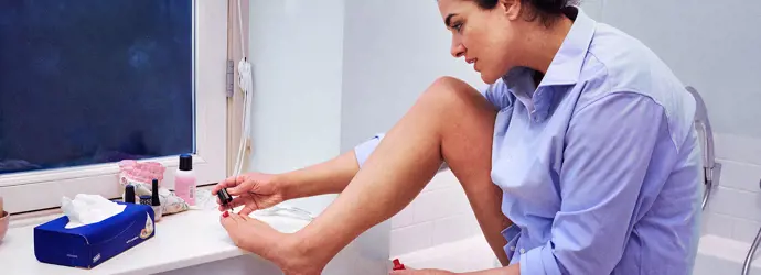 Una donna si sta dipingendo le unghie dei piedi in un bagno; davanti a lei c'è una scatola di fazzoletti.