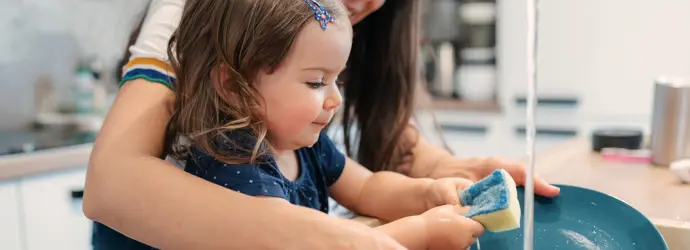 Una bambina che lava i piatti con l'aiuto di una donna sorridente.