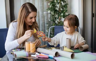 Una madre e una figlia fanno dei lavoretti insieme, utilizzando materiali riciclati come tubi di cartone e cartoncini.