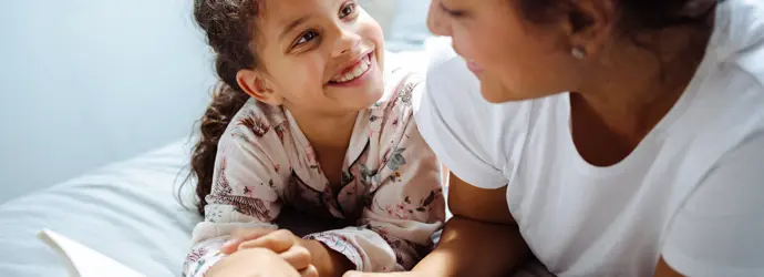 Una bambina e una donna sorridenti leggono insieme un libro su un letto.