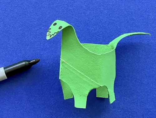 Dinosauro verde realizzato con rotolo di carta con occhietti neri su sfondo blu con accanto un pennarello nero.