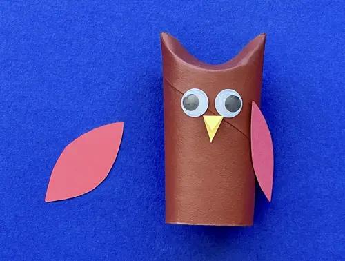 Rotolo di carta a forma di gufo marrone con occhietti, becco e un'ala. A sinistra l'ala mancante poggiata su sfondo blu.
