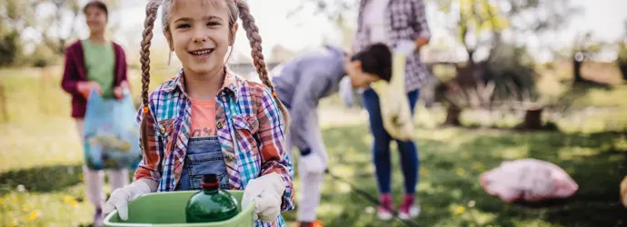 Una bambina con lunghe trecce tiene in mano un secchio verde, mentre sullo sfondo la sua famiglia raccoglie rifiuti da terra.