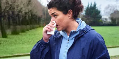 Eine Frau in einer dunkelblauen Jacke hält sich in einem Park ein Tempo Taschentuch vor ihre laufende Nase.
