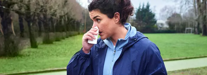 Eine Frau in einer dunkelblauen Jacke hält sich in einem Park ein Tempo Taschentuch vor ihre laufende Nase.
