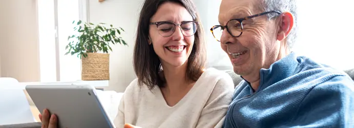 Una donna e un signore più anziano ridono mentre guardano qualcosa su un tablet seduti su un divano.