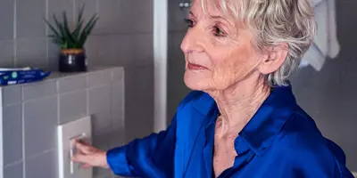 Eine ältere Frau in einer blauen Bluse betätigt die Spülung einer Toilette in einem gefliesten Badezimmer.