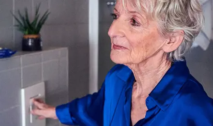 Eine ältere Frau in einer blauen Bluse betätigt die Spülung einer Toilette in einem gefliesten Badezimmer.
