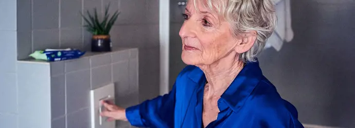 Eine ältere Frau in einer blauen Bluse betätigt die Spülung einer Toilette in einem gefliesten Badezimmer.
