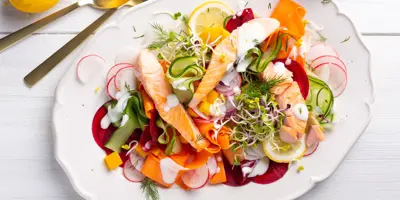 Bunter Salat aus Gurken, Möhren, Radieschen und Kräutern mit Lachs auf einem Teller garniert. Daneben goldenes Besteck und eine Zitrone.