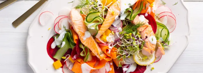 Bunter Salat aus Gurken, Möhren, Radieschen und Kräutern mit Lachs auf einem Teller garniert. Daneben goldenes Besteck und eine Zitrone.