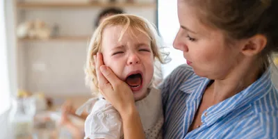 Eine Frau in blau-weiß gestreifter Bluse hält ihr weinendes Kleinkind im Arm und tröstet es nach einem Wutanfall.