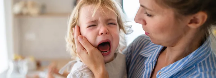 Eine Frau in blau-weiß gestreifter Bluse hält ihr weinendes Kleinkind im Arm und tröstet es nach einem Wutanfall.