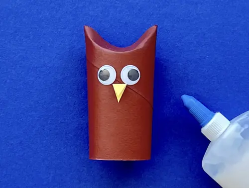 Rotolo di carta a forma di gufo color marrone con occhietti e becco su sfondo blu. Accanto un dispenser di colla bianco.